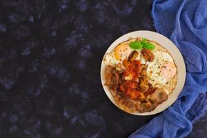 Fried eggs with bacon, tomatoes and mushrooms on dark background. Top view photo