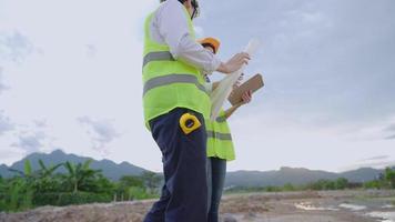 two Asian engineer construction workers walk and discuss on the work project, surveying looking on the blueprint paper and check on small detail using digital tablet, teamwork brainstorm cooperating video