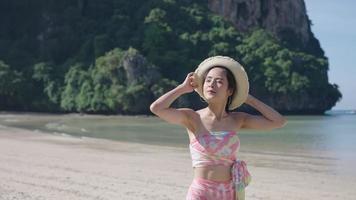 joven y bonita mujer asiática divirtiéndose caminando por la isla con el viento soplando, viaje de viaje en verano, mujer asiática con sombrero de paja disfrutando de las vacaciones de verano, clima cálido y soleado video