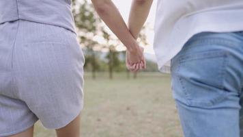 Rear view shot on a cheerful young couple hold hands while walking inside a summer grass field, closeup a swinging motions of hands with a tall green trees behind, relaxing exercise, side by side