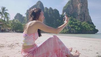 sidoporträtt en ung vacker asiatisk kvinna ringer videosamtal på smartphone, pratar glatt med att vifta med handen till en ny vän, kvinnlig soloresa på en strandresa till en tropisk ö, trådlöst nätverk video