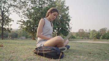 joven asiática sentada sola con una laptop en el parque verde, trabajando con una laptop en la escena al aire libre, campo de hierba verde, progresando en el proyecto escolar, autodedicación, escribiendo ensayo video