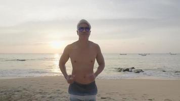 un anciano asiático activo corre desde la playa, un hombre de pelo gris fuerte y musculoso que usa gafas de sol corre en la playa disfruta del amanecer, entrenamiento de ejercicio remoto en la costa del mar de la isla, retiro de verano