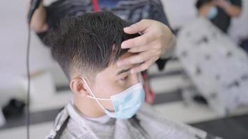 Asian man wear protective mask getting a hair cut during pandemic, reopening business, professional hairdresser drying client's hairs after cutting service, grooming check, new normal adaptation