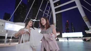 Attractive happy Asian women looking inside friend's shopping bag and cheerfully laughing together, fashionable females walking on modern pedestrian bridge in evening time, female hanging out
