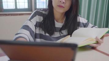 mulher asiática atraente sentada em casa trabalhando na mesa tocando no tablet com a outra mão segurando o livro didático, adolescente ativa fazendo autoestudo ou pesquisando sozinho na aconchegante sala de estar, aprendizagem online video