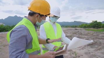 Asian male construction worker wear safety vest and hard helmet planning on building project, teamwork colleague, digital tablet blueprints paper, engineer and architect wear mask working together video