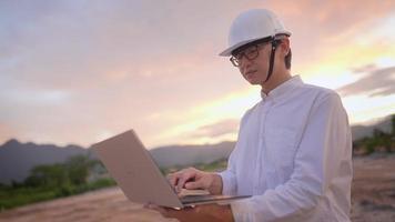 junger asiatischer ingenieur trägt brille, schutzhelm, der bei sonnenuntergang mit tragbarem laptop auf bauland im freien arbeitet, ingenieurlizenz, strukturplanungsanalysedesign, hart arbeitender mann