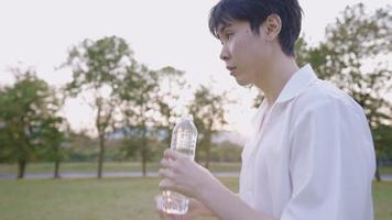 Young man drinking water from transparent plastic bottle standing in the park with sunset light on the background, human body temperature, healthy moisturizing skin, stay hydrate rehydration