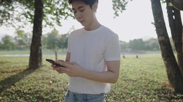 Young serious asian man using smartphone walking under trees inside green park on a hot sunny day, head down scrolling phone, portable device, mobile phone and modern technology, relaxing exercise video