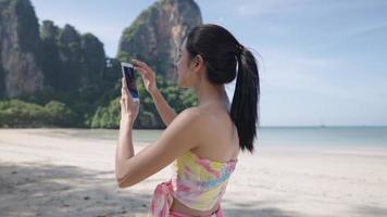 Young asian woman use cellphone taking picture while traveling, tropical island beach, wireless network connection. travel asia paradise island, beautiful summer beach. New gadget technology video