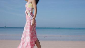 Young slim beautiful woman walking along the beach with a ocean and horizon sky background. Lady on vacation at tropical island. Wind blowing pink beach clothes, island shore fresh air, slow motion