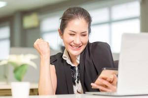 Beautiful Asian girl celebrate with laptop, Overjoyed woman looking at phone screen, celebrating success, showing yes gesture, Excited pretty girl using smartphone and celebrating success in office photo