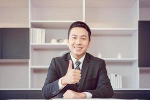 Smiling businessman sitting at office desk with showing thumbs up and looking at camera photo