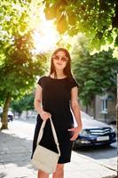 chica de negocios morena con vestido negro con gafas de sol con bolso a mano posando en la calle de la ciudad, coche de fondo en la carretera. foto