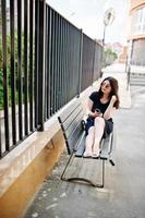chica morena con vestido negro, gafas de sol sentada en un banco, escuchando música desde el teléfono con auriculares y posando en la calle de la ciudad. foto
