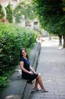 chica morena de negocios vestida de negro con gafas de sol sentada en la frontera contra arbustos, posando en la calle de la ciudad. foto