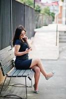 Brunette girl at black dress, sunglasses sitting on bench, listening music from headphones phone, and posing at street of city. photo