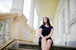 Brunette girl at black dress, sunglasses sitting on stairs of old vintage house, posing at street of city. photo