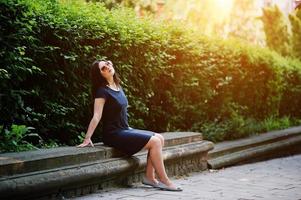 Brunette business girl at black dress on sunglasses sitting at border against bushes, posing at street of city. photo