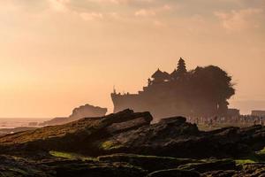 Ancient Tanah Lot temple with tourists on coastline in sunset photo