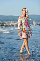 mujer rubia con vestido de pie en la playa de arena en la orilla del mar disfrutando de la vista de la puesta de sol. foto