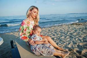 Mother and beautiful daughter having fun on the beach, sitting on sunbed. Portrait of happy woman with cute little girl on vacation. photo