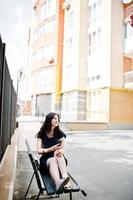 Brunette girl at black dress, sunglasses sitting on bench, listening music from headphones phone, and posing at street of city. photo