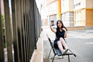 Brunette girl at black dress, sunglasses sitting on bench, listening music from headphones phone, and posing at street of city. photo