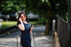 Brunette girl at black dress, sunglasses  listening music from headphones phone, and posing at street of city. photo
