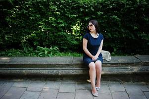 Brunette business girl at black dress on sunglasses sitting at border against bushes, posing at street of city. photo