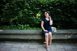 Brunette business girl at black dress on sunglasses sitting at border against bushes, posing at street of city. photo