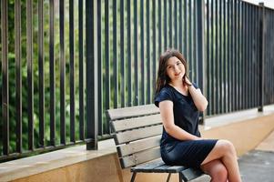 Brunette girl at black dress sitting on bench and posing at street of city. photo