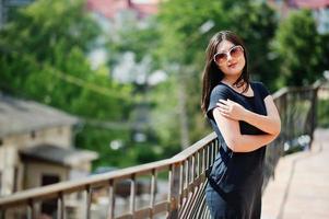 Brunette girl at black dress, sunglasses posing at street of city. photo