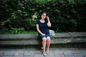 Brunette business girl at black dress on sunglasses sitting at border against bushes, posing at street of city. photo