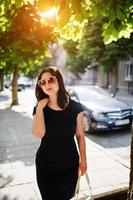 Brunette business girl at black dress on sunglasses with handbag at hand posing at street of city, background car on road. photo