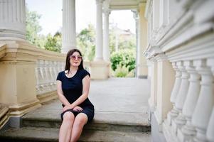 Brunette girl at black dress, sunglasses sitting on stairs of old vintage house, posing at street of city. photo