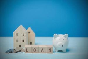 Concept saves money for loan buy house to the future, property. A white piggy bank LOAN on block wood with stack coins and wood house models on an old wooden table blue background with copy space. photo