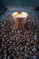 copper cup filled with espresso coffee in center of raw coffee beans spread out on rustic table photo