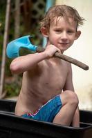 young boy playing in mud in wheelbarrow with shovel filled with water