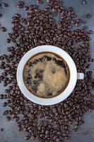 white coffee cup filled with strong coffee in center of raw coffee beans spread out on rustic table flat lay