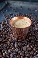 taza de cobre llena de café expreso en el centro de granos de café crudos esparcidos sobre una mesa rústica