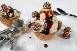 vanilla ice cream scoops with colorful candy pieces and chocolate syrup in waffle cone bowl on rustic white background photo