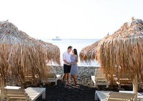 A guy and a girl look into each other's eyes against the background of the sea. In front of them are bungalows. Shot on Santorini Beach. photo