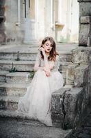 Bride.Young fashion model with perfect skin and makeup, white background. Beautiful bride on background white stairs. A woman in a long white dress is sitting on the stairs. photo