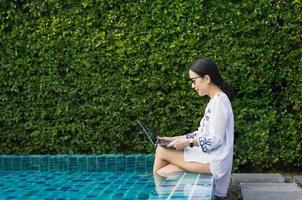 Woman working on laptop computer sitting at poolside on vacation. photo