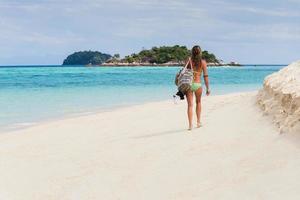 mujer en bikini con bolsa de playa caminando por la playa en isla tropical. foto