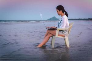 mujer de negocios de vista lateral usando una computadora portátil mientras está sentada en un chaie en la playa. foto