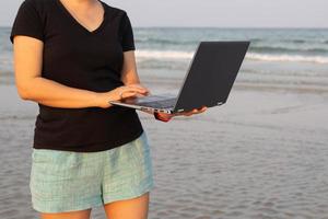 mujer de pie en la playa sosteniendo una computadora portátil en busca de señal. foto