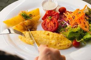 Woman hand holding fork and knife eating breakfast with omelet potato, tomatoes and vegetable salad on table. photo